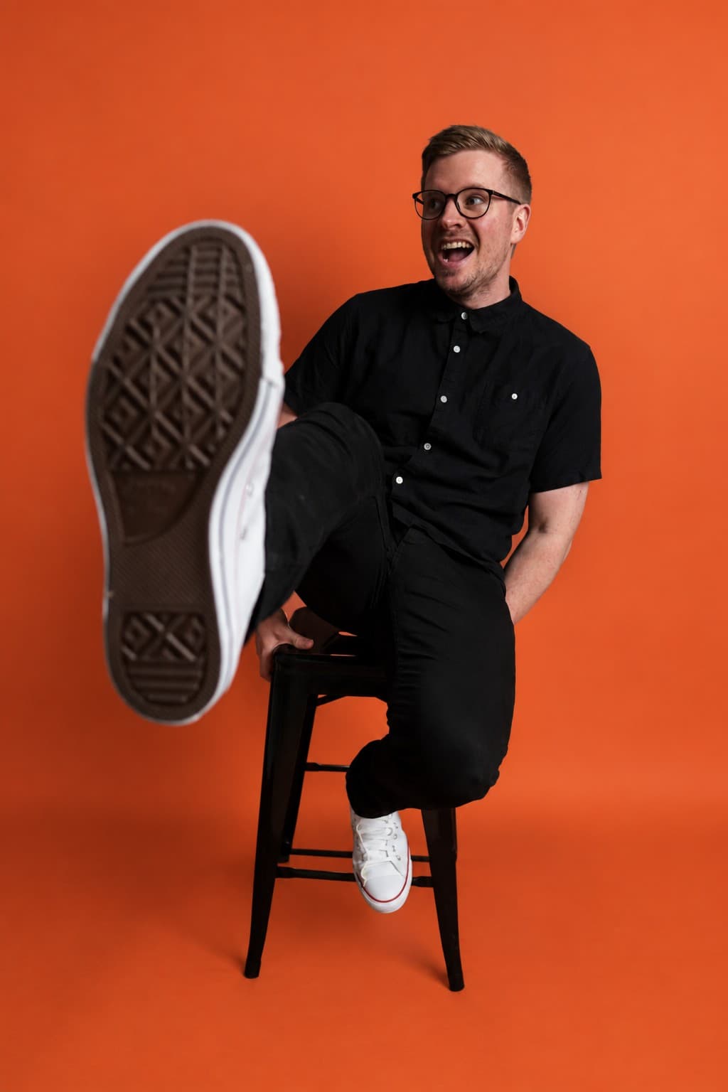 Shane Michael Hatton laughing on a stool, one leg kicked out, on an orange background