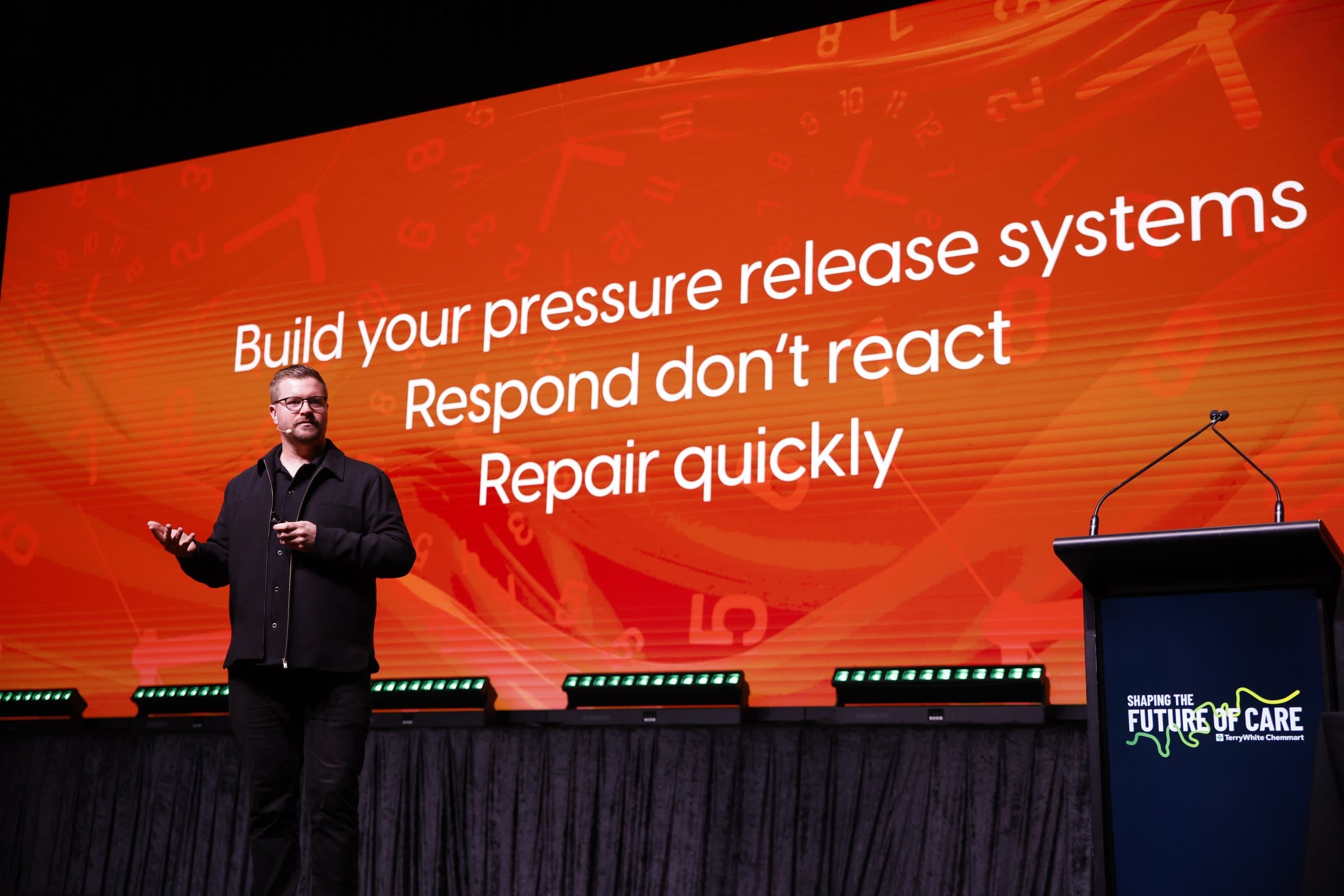Shane Michael Hatton on stage, speaking in front of an orange slide